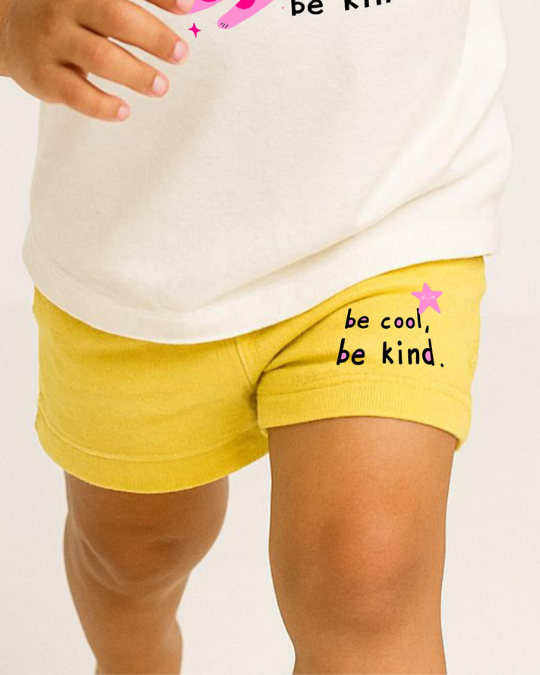 Person wearing a white shirt and yellow shorts with 'be cool, be kind.' text on a plain background