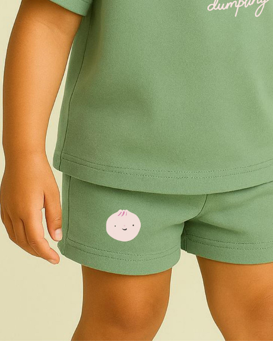 Green children's outfit with a small pink character on shorts against a beige background