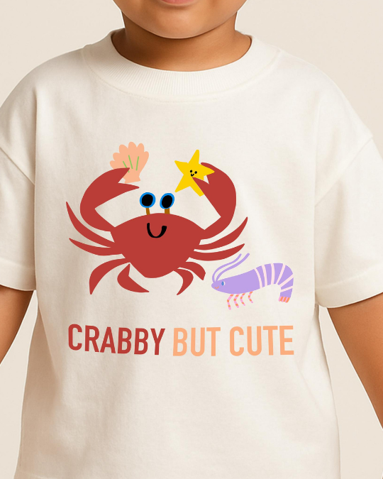 Child wearing a white t-shirt with a red crab and purple shrimp design, and 'CRABBY BUT CUTE' text.