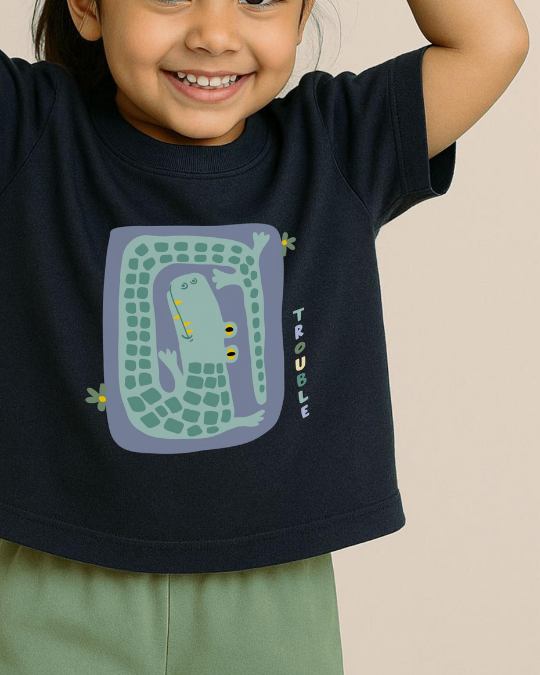 Child wearing a navy blue t-shirt with a crocodile graphic and 'Trouble' text on a beige background