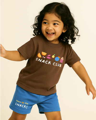 Child wearing a brown 'SNACK CLUB' t-shirt and blue shorts with text on a beige background